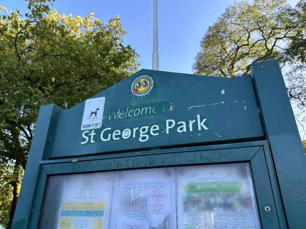 A green park sign reading "Welcome to St George Park," surrounded by trees with a blue sky in the background.